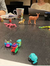 na zdjęciu widoczny stół z leżącymi kolorowymi figurkami zwierząt, za stołem widoczne fragmenty dwóch siedzących postaci