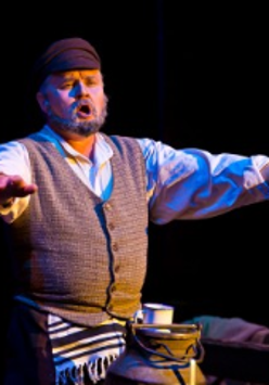 pośrodku zdjęcia mężczyzna siedzący z wyciągniętymi w bok obiema rękami, przed nim widoczna górna część dużej kanki na mleko, na niej kubek; mężczyzna śpiewa.