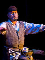 pośrodku zdjęcia mężczyzna siedzący z wyciągniętymi w bok obiema rękami, przed nim widoczna górna część dużej kanki na mleko, na niej kubek; mężczyzna śpiewa.