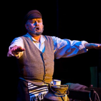 pośrodku zdjęcia mężczyzna siedzący z wyciągniętymi w bok obiema rękami, przed nim widoczna górna część dużej kanki na mleko, na niej kubek; mężczyzna śpiewa.