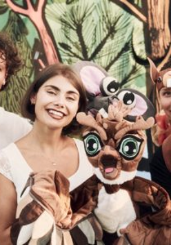 Zdjęcie trójki aktorów, którzy trzymają lalki - bohaterów spektaklu Grajmy w zielone. Aktorzy stoją na tle rysunkowego lasu.