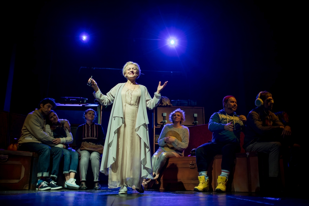 Grupa aktorów siedzi na scenie. Na środku stoi kobieta, ma ręce do góry, w ręce dzwonek. Uśmiecha się. Ubrana jest w jasny kostium.