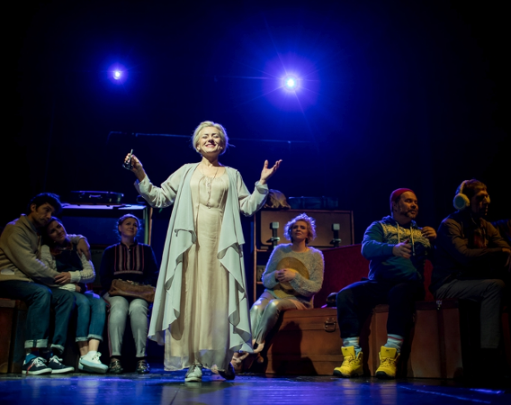 Grupa aktorów siedzi na scenie. Na środku stoi kobieta, ma ręce do góry, w ręce dzwonek. Uśmiecha się. Ubrana jest w jasny kostium.