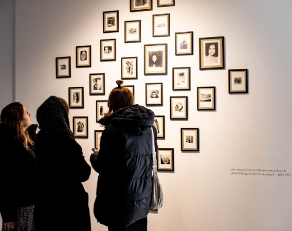 Grupa ludzi przygląda się wielu małym fotografiom wiszącym na białej ścianie.