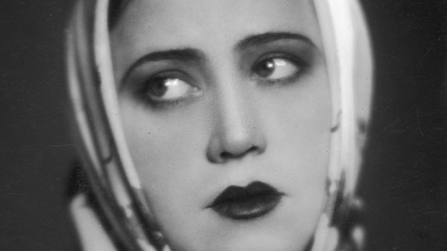 Czarno-biały portret młodej kobiety z chustką na głowie. Kobieta ma mocny makijaż. Patrzy nieco w bok, ma rozmarzone spojrzenie.