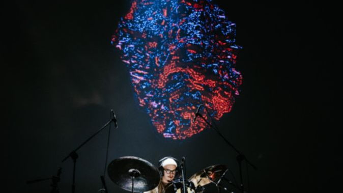 Perkusista w słuchawkach na uszach gra na perkusji. Za nim czerwono-niebieska wizualizacja przypominająca głaz.