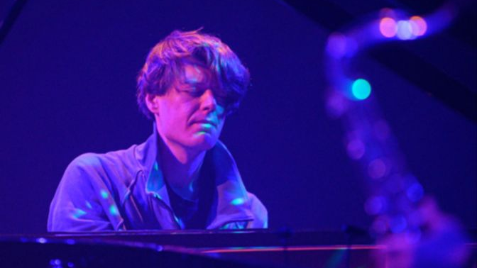 Mężczyzna z zamkniętymi oczami gra na fortepianie skąpany w różowo-niebieskim świetle.