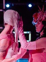 Na ciemnym tle dwie postaci w różowych strojach, stoją bokiem do widza, przodem do siebie.