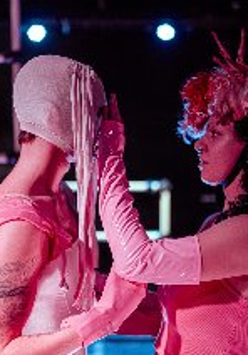 Na ciemnym tle dwie postaci w różowych strojach, stoją bokiem do widza, przodem do siebie.