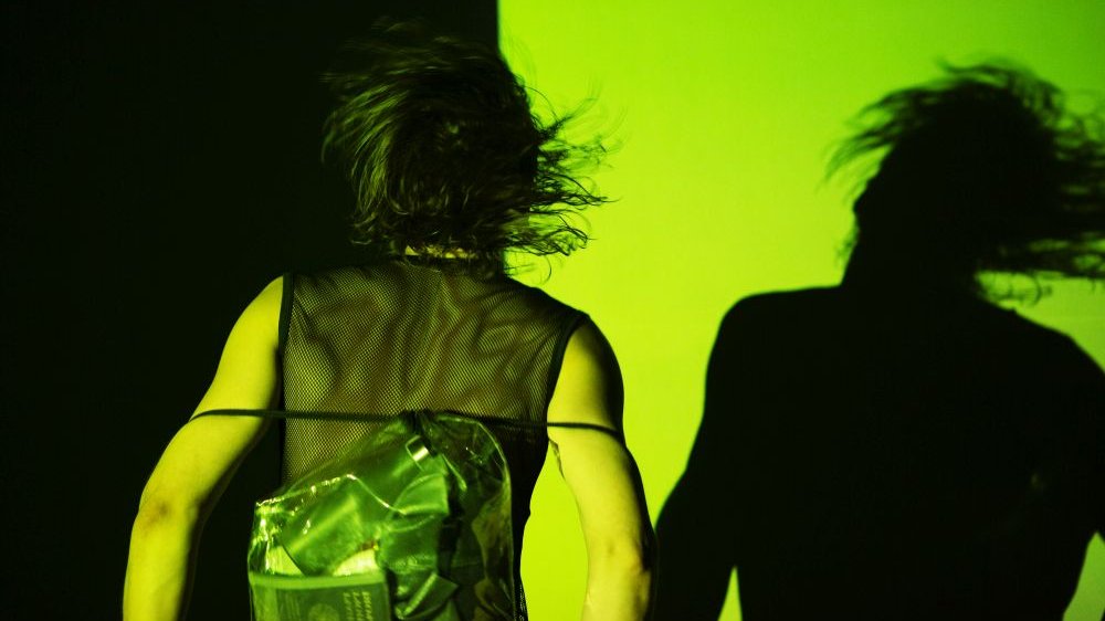 Tancerz odwrócony tyłem do obiektywu, w trakcie ruchu, jest ubrany w siatkowaną koszulkę. Rzuca wykrzywiony cień na ścianę w kolorze jadowitej zieleni.