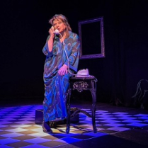 Zdjęcie aktorki na scenie, opartej o stolik, rozmawiającej przez telefon.