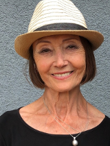 Ewa Wydyłło. Porter kobiety w średnim wieku. Kobieta ma krótkie, brązowe włosy, na głowie kapelusz. Patrzy przed siebie i szeroko się uśmiecha.