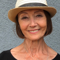 Ewa Wydyłło. Porter kobiety w średnim wieku. Kobieta ma krótkie, brązowe włosy, na głowie kapelusz. Patrzy przed siebie i szeroko się uśmiecha.