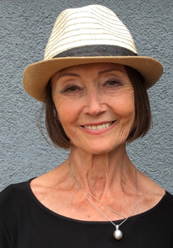 Ewa Wydyłło. Porter kobiety w średnim wieku. Kobieta ma krótkie, brązowe włosy, na głowie kapelusz. Patrzy przed siebie i szeroko się uśmiecha.