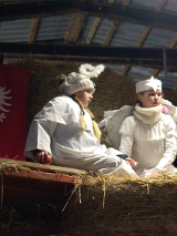 Dwie dziewczynki w strojach aniołów siedzą na belce w stodole, za nimi widać zawieszoną na ścianie czerwoną flagę z białym orłem.