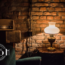 Stara miedziana lampa z ozdobnym kloszem stoi na stoliku. Oświetla zielony fotel, rozwieszoną gazetę i stare radio. W tle ceglana ściana.