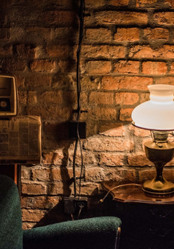 Stara miedziana lampa z ozdobnym kloszem stoi na stoliku. Oświetla zielony fotel, rozwieszoną gazetę i stare radio. W tle ceglana ściana.