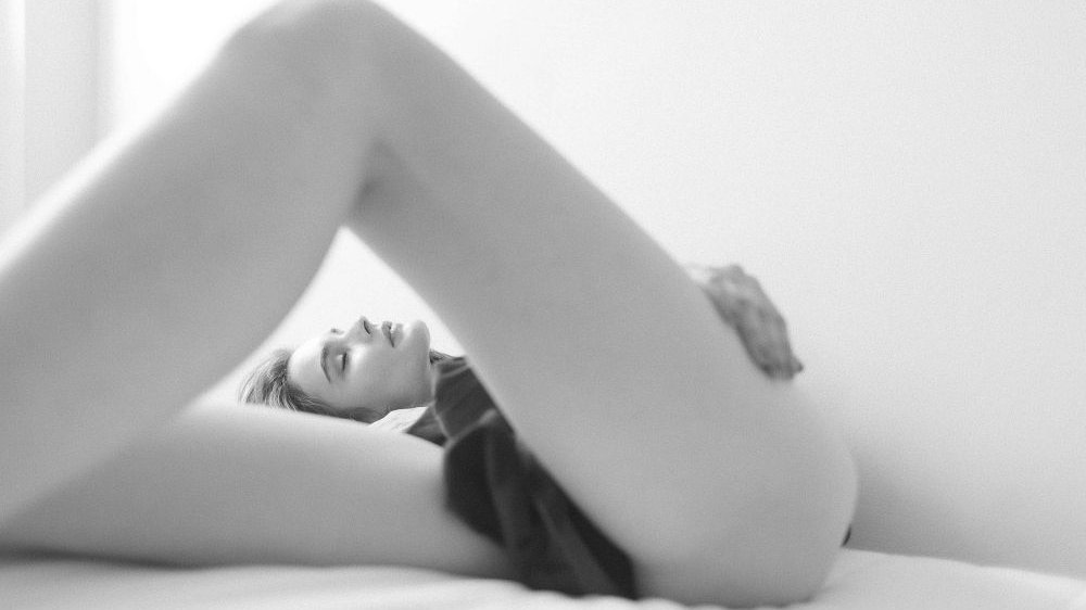 Czarno-biała fotografia modelki leżącej na łóżku. Na pierwszym planie jej nogi.