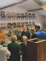 Obraz przedstawia grupę ludzi uważnie dyskutujących lub badających jakiś obiekt w warsztacie lub muzeum. Pokój wydaje się być warsztatem lub wycieczką. Ściany ozdobione są rzędami krzeseł o różnych wzorach i kolorach, podkreślając temat miejsca, być może związany z projektowaniem mebli lub rzemiosłem. Przestrzeń ma nieco antyczny wygląd z widocznymi drewnianymi belkami i tynkiem na ścianach. Oświetlenie jest miękkie, tworząc przytulną atmosferę.