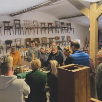 Obraz przedstawia grupę ludzi uważnie dyskutujących lub badających jakiś obiekt w warsztacie lub muzeum. Pokój wydaje się być warsztatem lub wycieczką. Ściany ozdobione są rzędami krzeseł o różnych wzorach i kolorach, podkreślając temat miejsca, być może związany z projektowaniem mebli lub rzemiosłem. Przestrzeń ma nieco antyczny wygląd z widocznymi drewnianymi belkami i tynkiem na ścianach. Oświetlenie jest miękkie, tworząc przytulną atmosferę.