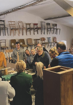 Obraz przedstawia grupę ludzi uważnie dyskutujących lub badających jakiś obiekt w warsztacie lub muzeum. Pokój wydaje się być warsztatem lub wycieczką. Ściany ozdobione są rzędami krzeseł o różnych wzorach i kolorach, podkreślając temat miejsca, być może związany z projektowaniem mebli lub rzemiosłem. Przestrzeń ma nieco antyczny wygląd z widocznymi drewnianymi belkami i tynkiem na ścianach. Oświetlenie jest miękkie, tworząc przytulną atmosferę.