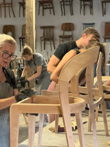 Na zdjęciu widać grupę osób pracujących nad drewnianymi krzesłami w warsztacie. Na obrazie znajduje się cztery osoby, wszystkie ubrane w codzienne ubrania i pracujące nad różnymi aspektami krzeseł. Krzesła są wykonane z jasnego drewna i mają prosty, nowoczesny design. W warsztacie rozrzucone są narzędzia i materiały, a ściany ozdobione są półkami z gotowymi krzesłami. Osoby na zdjęciu są skoncentrowane na swojej pracy, a atmosfera jest pełna twórczości i rzemiosła.