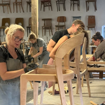 Na zdjęciu widać grupę osób pracujących nad drewnianymi krzesłami w warsztacie. Na obrazie znajduje się cztery osoby, wszystkie ubrane w codzienne ubrania i pracujące nad różnymi aspektami krzeseł. Krzesła są wykonane z jasnego drewna i mają prosty, nowoczesny design. W warsztacie rozrzucone są narzędzia i materiały, a ściany ozdobione są półkami z gotowymi krzesłami. Osoby na zdjęciu są skoncentrowane na swojej pracy, a atmosfera jest pełna twórczości i rzemiosła.