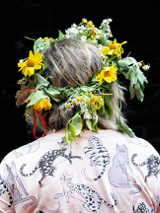 Osoba odwrócona tyłem. Na głowie ma wianek z żółtych i polnych kwiatów. Na sobie ma różową koszulę z printem w koty.