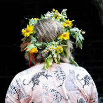 Osoba odwrócona tyłem. Na głowie ma wianek z żółtych i polnych kwiatów. Na sobie ma różową koszulę z printem w koty.