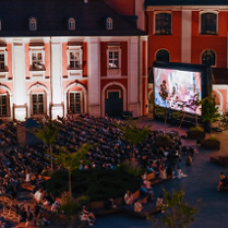 Widok z góry na plac Kolegiacki, na którym stoi duży ekran kinowy i rozłożone leżaki. Na ekranie wyświetla się film. Na leżakach siedzi widownia. Wokół małe drzewka. W tle budynek Kolegiaty.