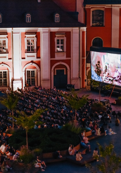 Widok z góry na plac Kolegiacki, na którym stoi duży ekran kinowy i rozłożone leżaki. Na ekranie wyświetla się film. Na leżakach siedzi widownia. Wokół małe drzewka. W tle budynek Kolegiaty.