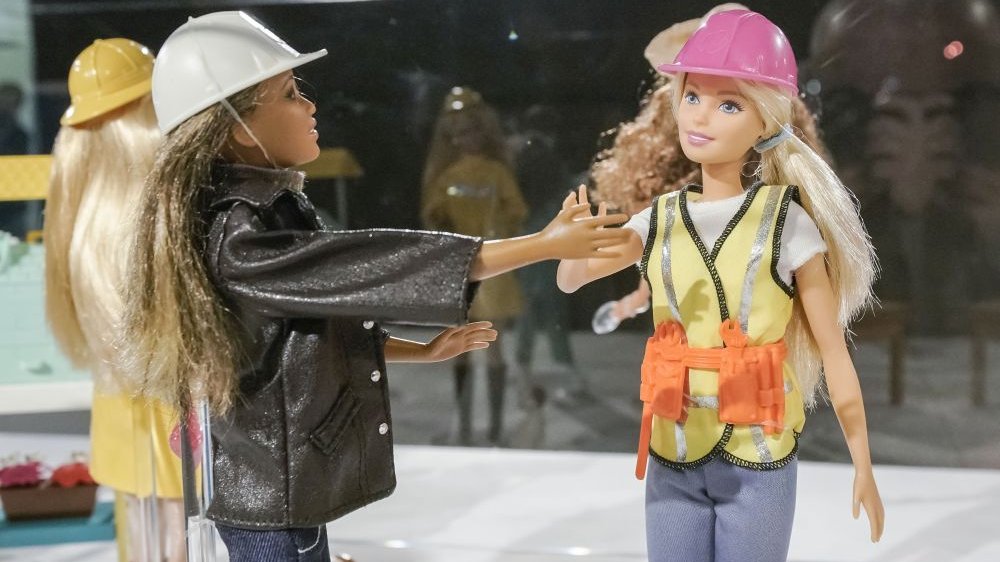 Na zdjęciu dwie lalki Barbie. Ta po prawej jest blondynką ubraną w kamizelkę roboczą, a na głowie ma różowy kask. Ta po lewej jest ciemnowłosą mulatką, ma szary kask.