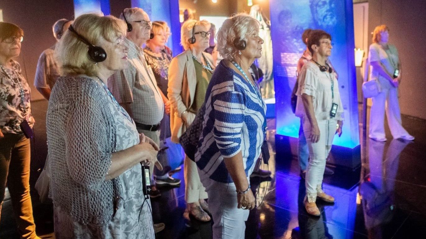 Grupa seniorów zwiedzających ekspozycję, wpatrzonych w jeden punkt, na głowach mają słuchawki, a na szyi zawieszone audioprzewodniki.