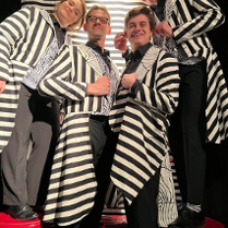 Zdjęcie aktorów - kobieta i trzech mężczyzn we frakach w biało-czarne paski. Stoją obok siebie, wpatrzeni w obiektyw, uśmiechają się.