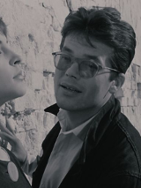 Czarno-biały kadr kobiety w słońcu opierającej się o ścianę, a obok niej mężczyzna w okularach.