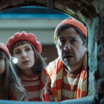 Dziewczynka, kobieta i mężczyzna ubrani w podobne stroje i czerwone nakrycia na głowie zaglądają do wąskiego pomieszczenia.