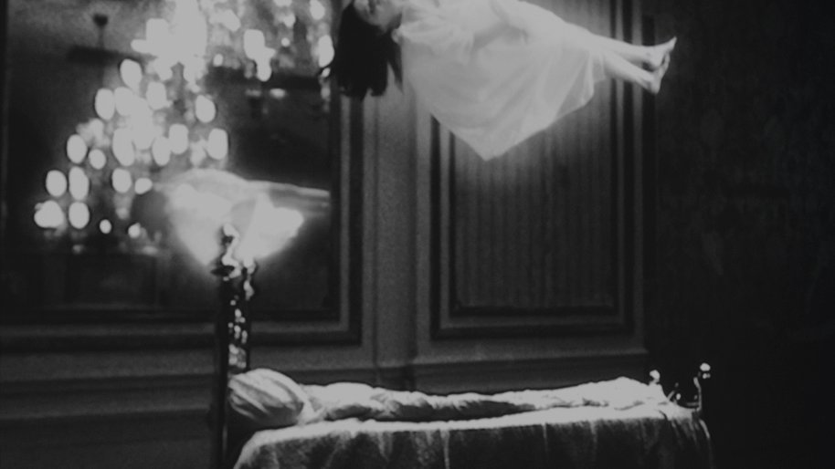 Łóżko, nad którym unosi się dziewczyna w białej sukni. Obok żyrandol. Czrano-białe zdjęcie.