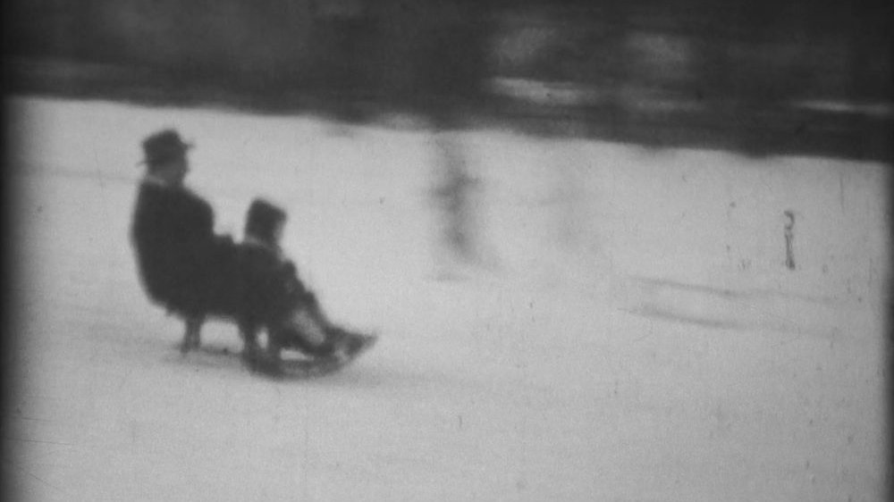 Niewyraźne zdjęcie będące fragmentem klatki filmowej. Na nim ojciec z dzieckiem zjeżdżający na sankach po śniegu z górki położonej w mieście.