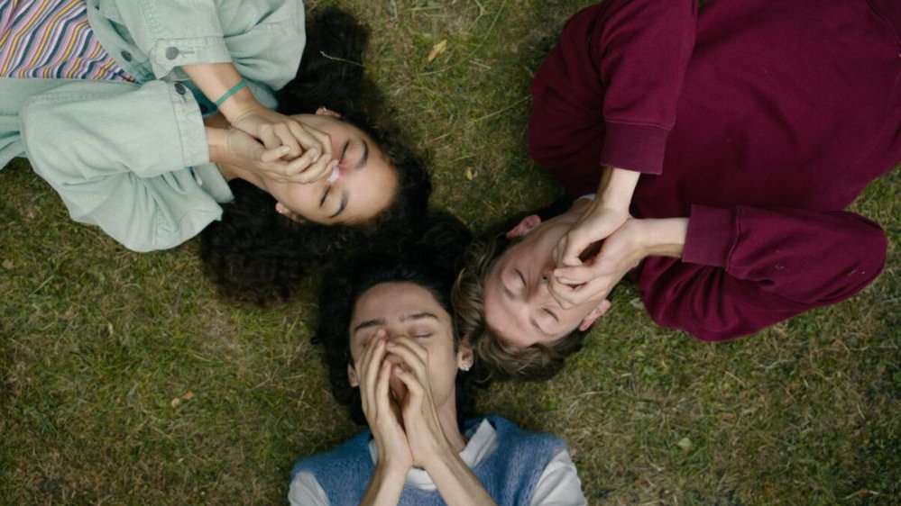 Troje młodych ludzi widzianych z lotu ptaka. Leżą na trawie, rękoma zasłaniają usta.