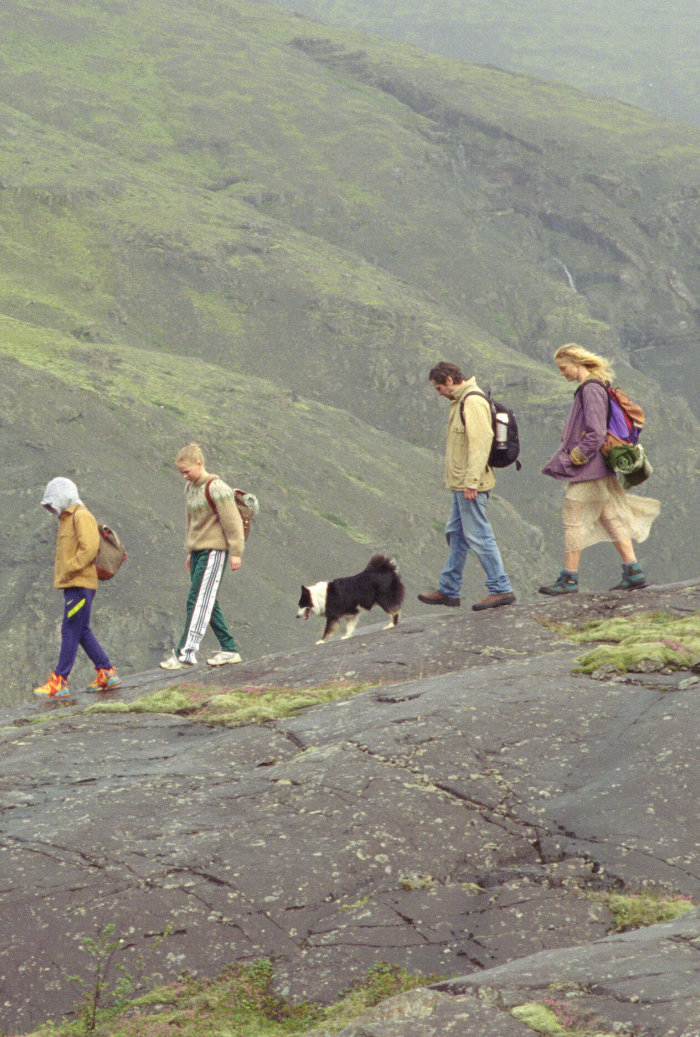 Kadr z filmu. Para dosrosłych i para dzieci idzie szlakiem w górach. Pomiędzy nimi wędruje pies. Wieje wiatr.