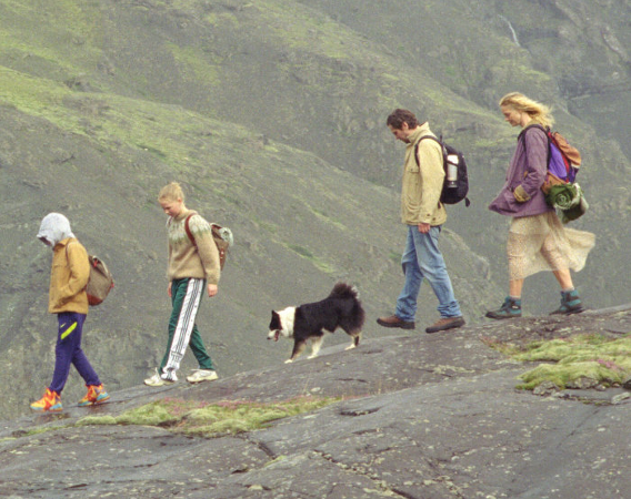 Kadr z filmu. Para dosrosłych i para dzieci idzie szlakiem w górach. Pomiędzy nimi wędruje pies. Wieje wiatr.