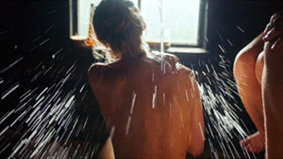 Kobieta odwrócona tyłem do obiektywu, bierze prysznic. Stoi przodem do okna.