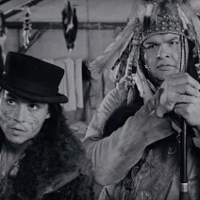 Dwoje mężczyzn. Po prawej Indianin - rdzenny amerykanin, po lewej mężczyzna z kapeluszu i skórze owczej. Za nimi ściana namiotu.