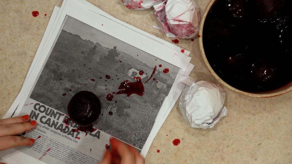 Zakrwawiona gazeta, dwie miski z krwią i kobiece dłonie z paznokciami pomalowanymi na czerwono.