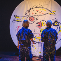 na zdjeciu widoczne dwie osoby stojące tyłem, w granatowych kombinezonach popisanych kolorowymi farbami, przed nimi biala płaszczyzna w ksztalcie koła, na której narysowano kolorowe ryby i ptaki