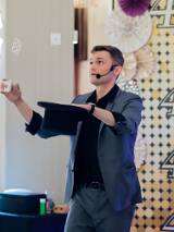 na zdjęciu mężczyzna w ciemnym garniturze, w jednej ręce trzyma czarny kapelusz, w drugiej talię kart