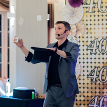 na zdjęciu mężczyzna w ciemnym garniturze, w jednej ręce trzyma czarny kapelusz, w drugiej talię kart