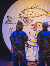 na zdjeciu widoczne dwie osoby stojące tyłem, w granatowych kombinezonach popisanych kolorowymi farbami, przed nimi biala płaszczyzna w ksztalcie koła, na której narysowano kolorowe ryby i ptaki