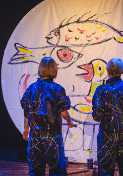 na zdjeciu widoczne dwie osoby stojące tyłem, w granatowych kombinezonach popisanych kolorowymi farbami, przed nimi biala płaszczyzna w ksztalcie koła, na której narysowano kolorowe ryby i ptaki