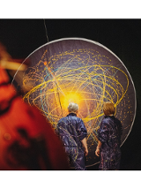 na zdjeciu widoczne dwie osoby stojące tyłem, w granatowych kombinezonach popisanych kolorowymi farbami, przed nimi czarna płaszczyzna w kształcie koła, na której narysowano kolorowe okręgi i elipsy